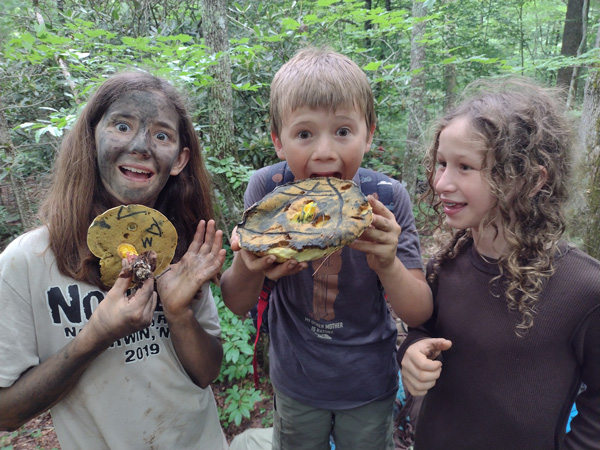 Children at Asheville forest school goofing around with large mushrooms wearing natural camouflage face paint