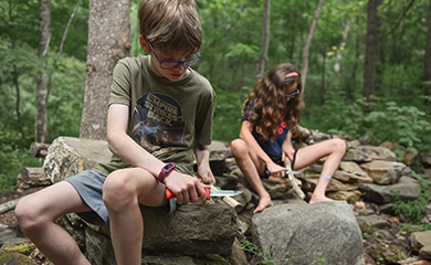 Children sitting on boulders in the forest carving sticks with knives at Asheville forest school for bushcraft