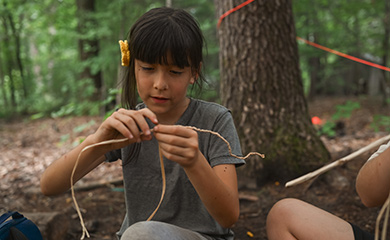 Young girl weaving cordage from natural materials at bushcraft program for Asheville forest homeschooling