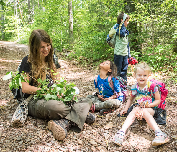 Nature connection mentor seated with children on a gravel path examining a fallen tree branch